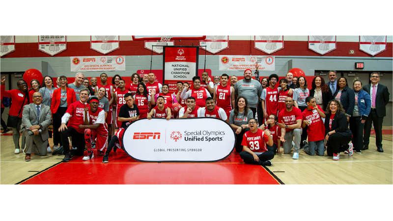 Students and teachers stand pose for a group photo in a high school gym. They are standing behind a sign that reads "ESPN Special Olympics Unified Sports Global Presenting Sponsor". 