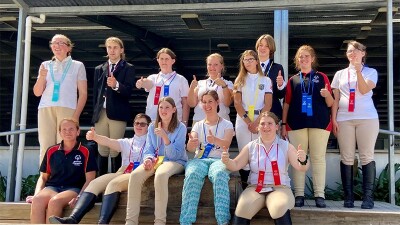 A group of equestrian athletes posing for a photo, some holding enthusiastic ‘thumbs up.’ Most have at least one ribbon around their neck.