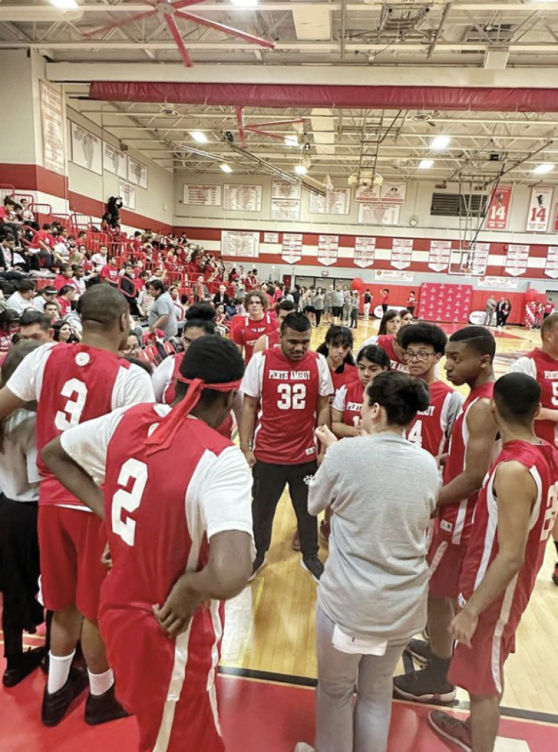 A basketball team huddles on the sidelines of a gymnasium. They are all wearing matching red and white uniforms and the gym is packed with fans. 