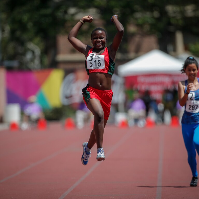 Three female runners on the track; female in the middle is jumping in celebration.