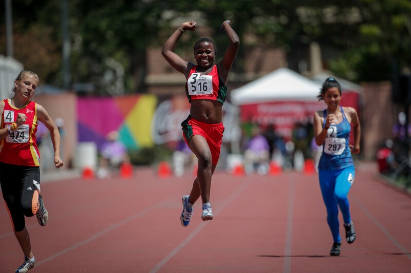 Three female runners on the track; female in the middle is jumping in celebration.