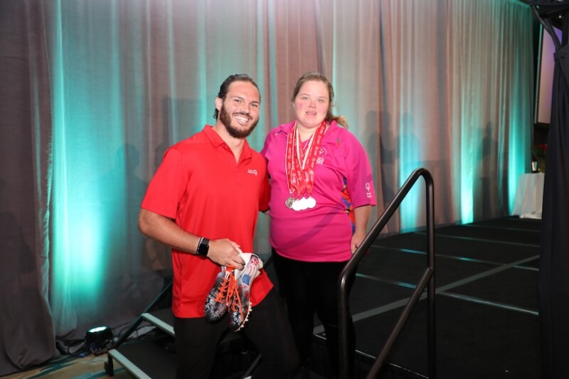 Alex Singleton, a NFL player, poses for a photo with his sister, Ashley, a Special Olympics athlete. Alex is holding his NFL cleats and Ashley is wearing her medals. 