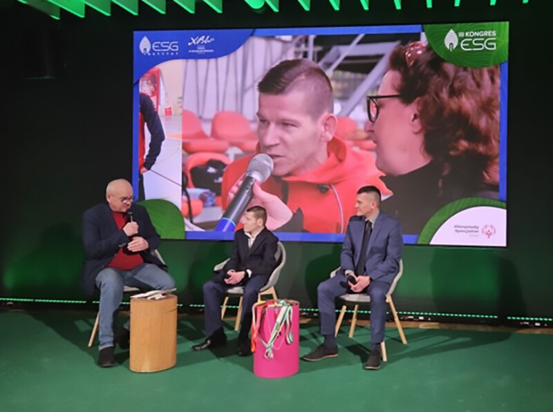 One man holding a microphone looks towards Konrad and another man sitting alongside him with a large screen of a man being interviewed in the background. 