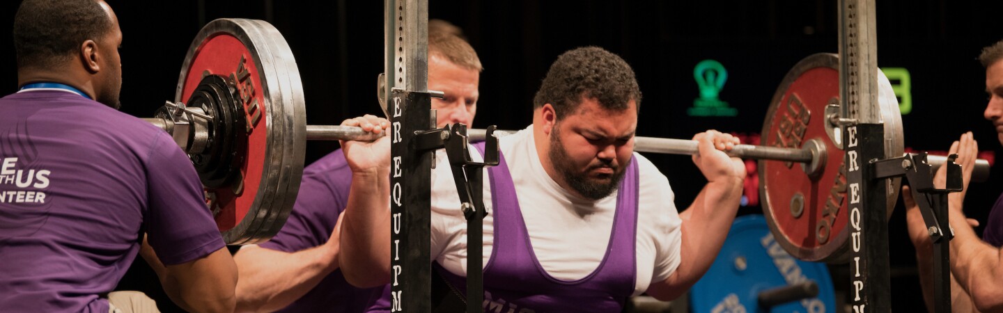 male lifter performs a squat at the rack while coaches and officials assist. 