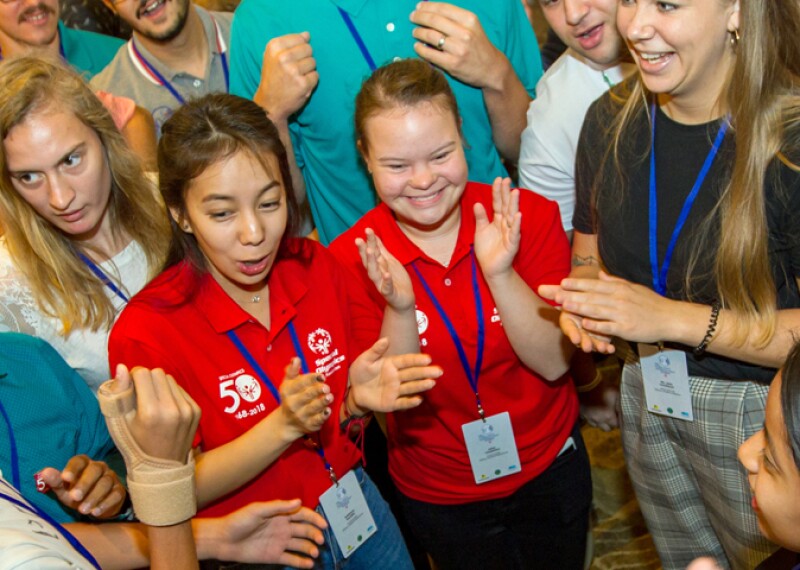 A large group of athletes and Special Olympics representatives stand in a circle clapping with joyful expressions on their faces. 