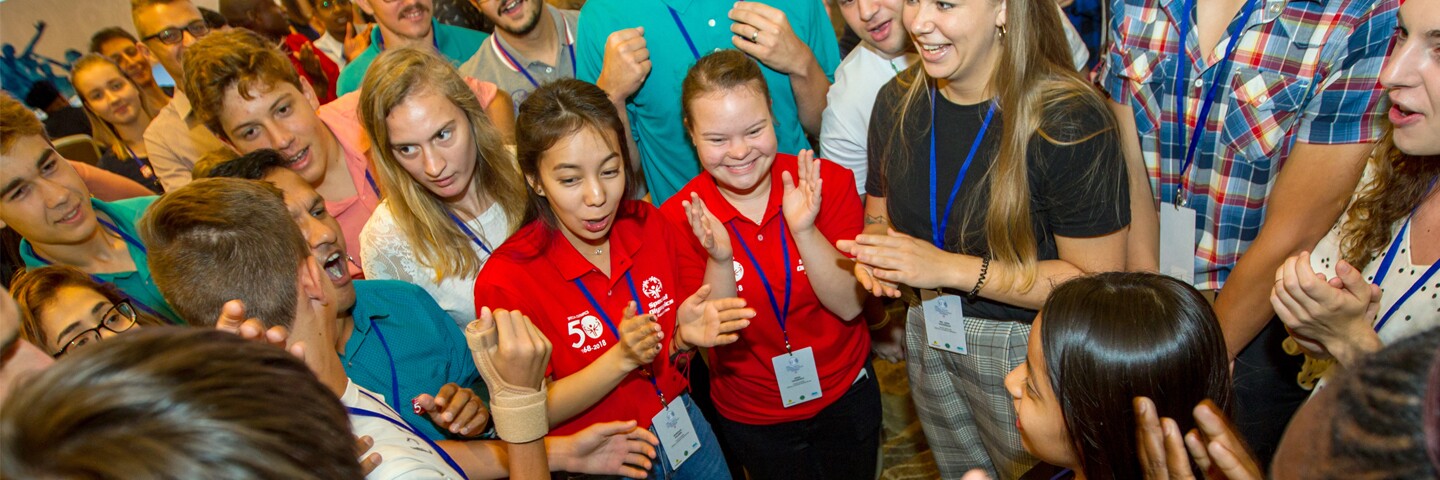 A large group of athletes and Special Olympics representatives stand in a circle clapping with joyful expressions on their faces. 