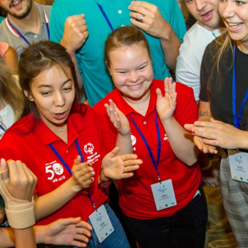 A large group of athletes and Special Olympics representatives stand in a circle clapping with joyful expressions on their faces. 