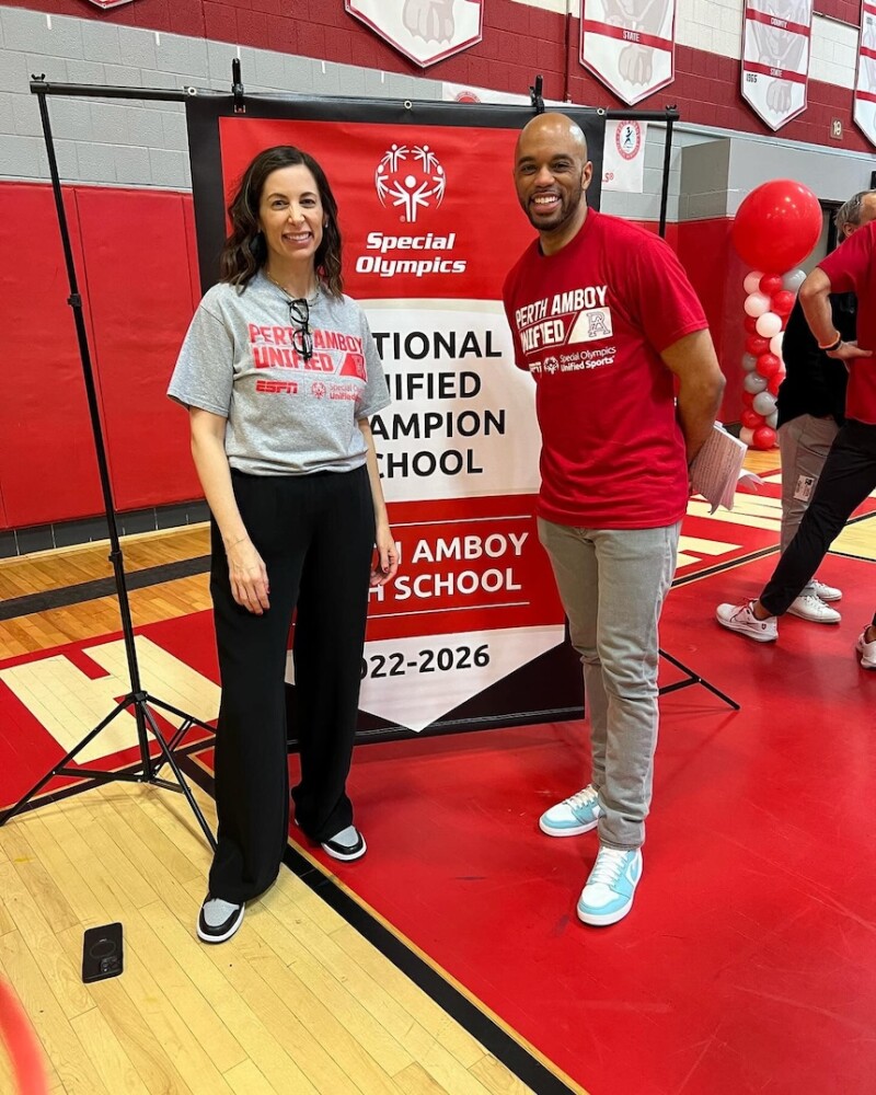 Two individuals stand in a gym in front of a banner that reads "Special Olympics National Unified Champion School Perth Amboy High School 2022-2026". 