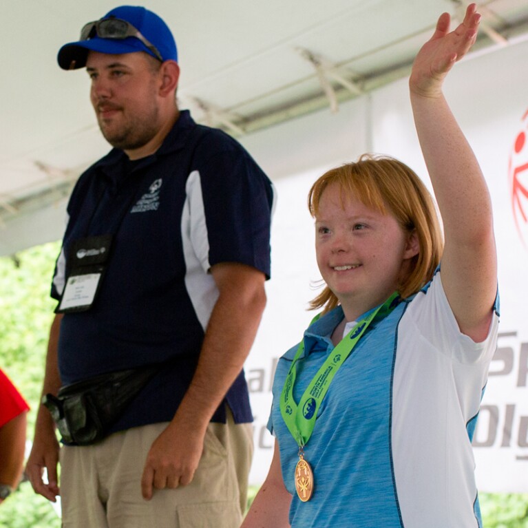 Young woman with a medal around her neck raising her arm and a young man standing next to her. 