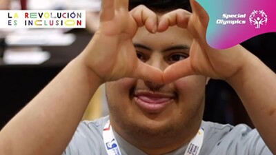 A Special Olympics athlete who got free health screenings in the Healthy Athletes Program during World Games Abu Dhabi 2019 makes a heart-shaped sign with his hands while looking at camera. Spanish text in front of him reads "Together, we have the power to make sure nobody is left behind during this critical moment."