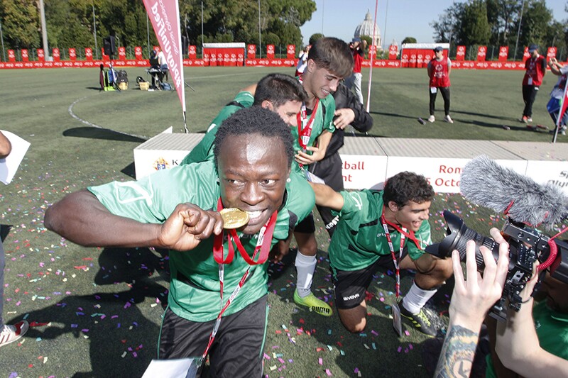 Young man on a football pitch, in front of a podium celebrating with four teammates and biting a golden medal