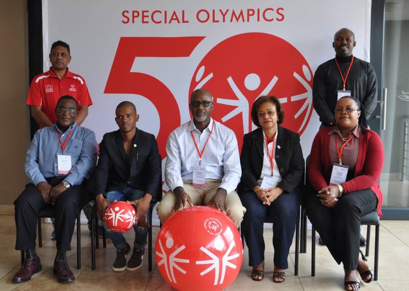 5 people sit and 2 people stand looking official in front of a Special Olympics 50th anniversary banner. 