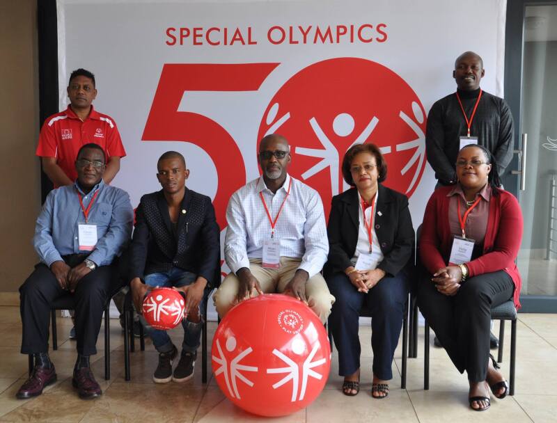 5 people sit and 2 people stand looking official in front of a Special Olympics 50th anniversary banner. 