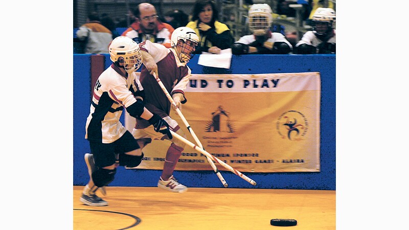 Athletes run toward the floor hockey puck while a member from the opposing team tries to block. 