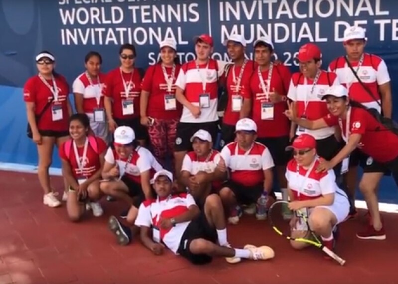 Athletes, representatives, and friends gather together in a group in front of the World Tennis Invitational sign for a group photo. Every is wearing a white and red or red polo shirt. 