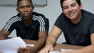 Exsaul and Rodrigo sit at a desk next to each other, each holding papers and smiling at the camera.