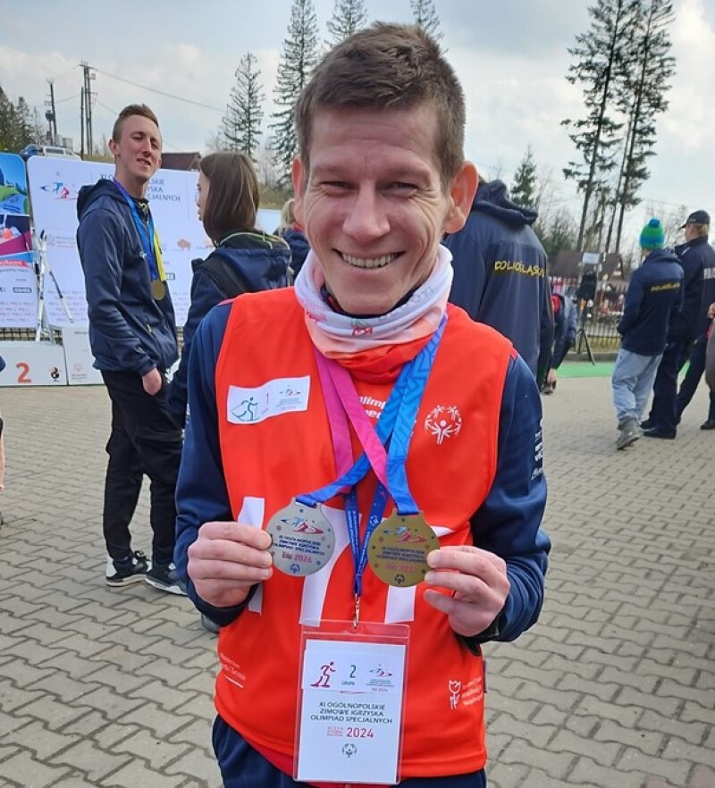 Konrad is wearing a red Special Olympics jersey and smiles at the camera while holding his medals.