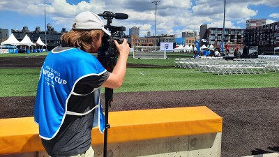 Man with blue media bib and white cap looks through a video camera on a tripod at a football-soccer field with a blue sky above dotted with white clouds.