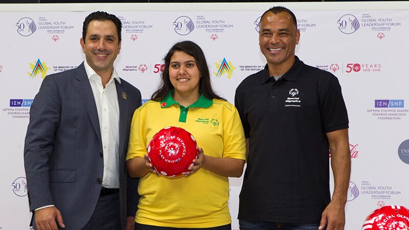 Three people standing side by side facing the camera. A man in a suit, a women holding a read ball and a man in a black Special Olympics t-shirt.