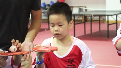 A young athlete is playing table tennis during 2018 Special Olympics East Asia Unified Camp. He is wearing a red and white Special Olympics jersey and a blue watch. He has a ping pong paddle in his right hand and balancing a ball on top which he is intently staring and consecrating on. 