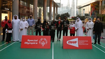 A group of people standing behind A-Board signage and a badminton court with large buildings in the backdrop.