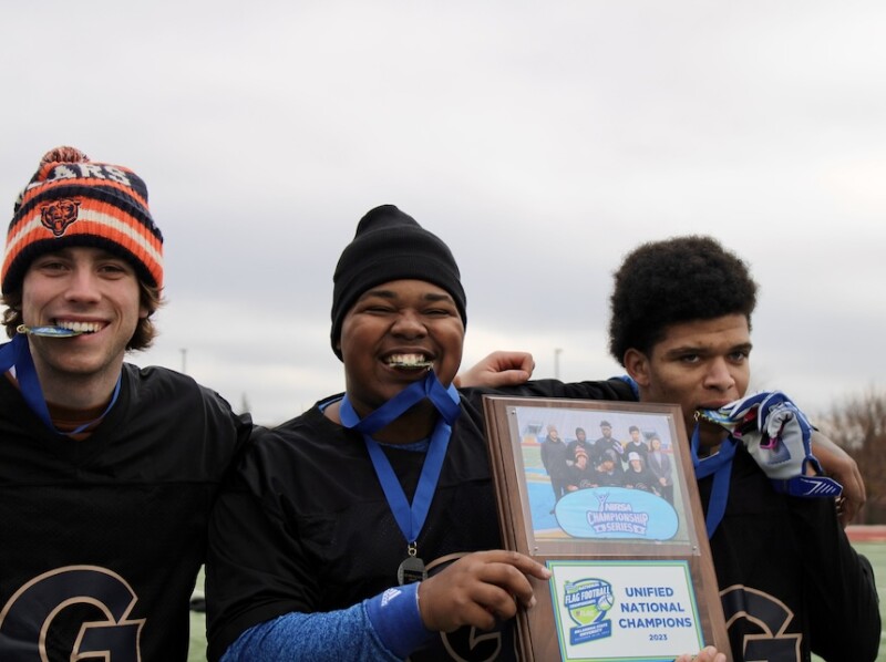 Three teammates pose for a photo. They're biting their medals and holding a plaque. 