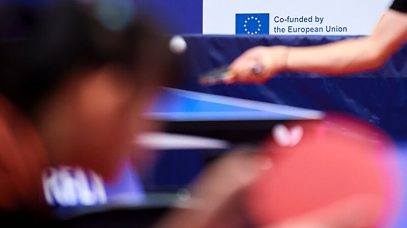 Table tennis players playing, the EU logo is in the background. 