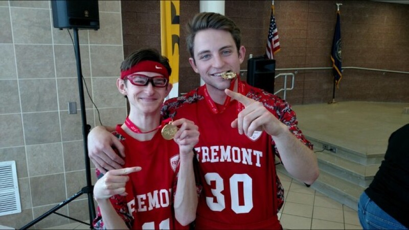 Two men wearing red basketball uniforms smile for a photo. They have their arms around each other and are wearing gold medals around their necks. 