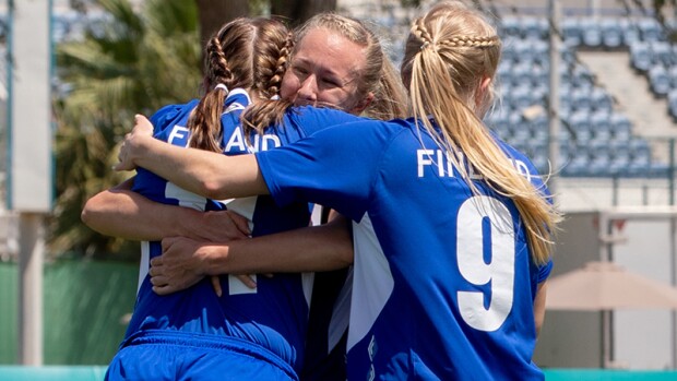 Three girls hug on the football pitch in celebration. 