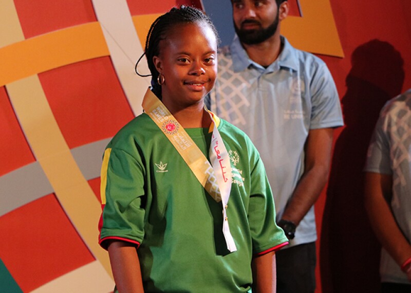 A smile from Team Senegal athlete Khadija Sy after the medal ceremony for the 100m run. She's standing on stage in front of the World Games logo with volunteers behind her. 