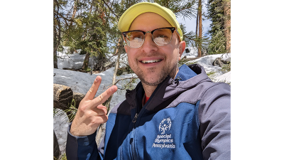 A man poses for a selfie. He's outside in front of a hill and is wearing a blue jacket with the Special Olympics Pennsylvania logo on it. 