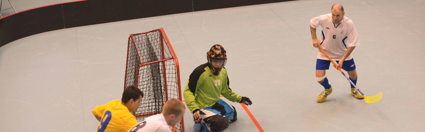 Athlete attempting to make a goal while his opponent blocks; the goalie and another player watch. 