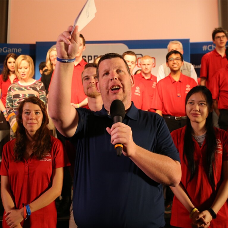 Man speaking into a microphone he's holding in his left hand and his right hand up holding note cards. Spectators are behind him. 