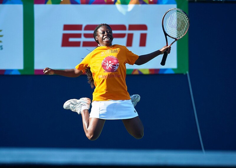 Female tennis athlete jumping up in the air for joy. She's holding her racquet in her left hand, has a white tennis skirt and a yellow Special Olympics t-shirt. 