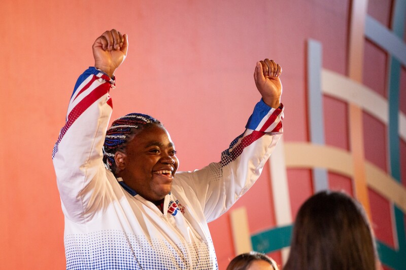 A Special Olympics athlete stands on top of the medal podium. She raises her arms above her head in celebration. 