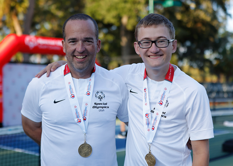 A father and son pose for a photo with their arms around each other. They are standing on a pickleball court and are wearing gold medals. 