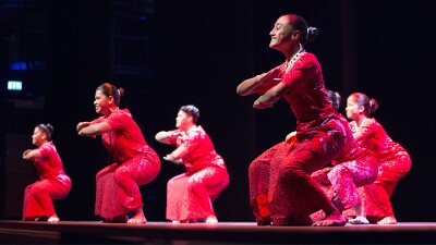 Six dancers on stage wearing red shirts and sarongs.