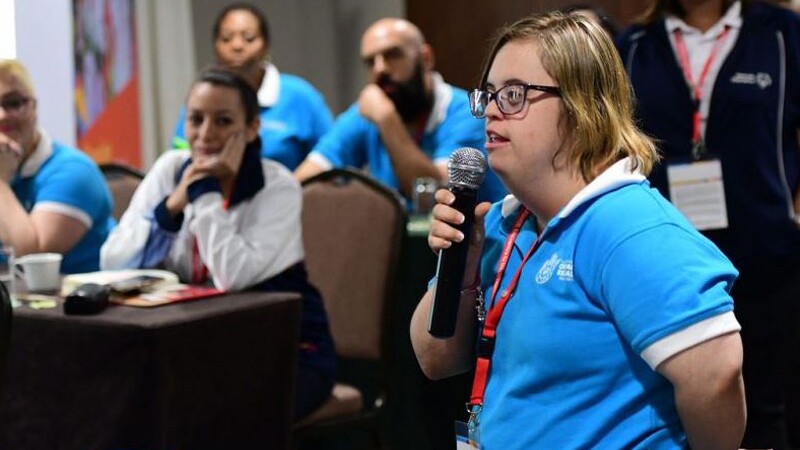 A female athlete leader holds a microphone as she talks at an event. People in the background listen to her.