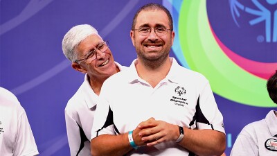 Man standing and smiling with his hands clasped and a man looking over his shoulder. 