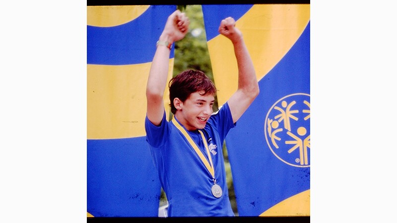 A male athlete with his arms in the air and a medal around his neck celebrates his medal win.