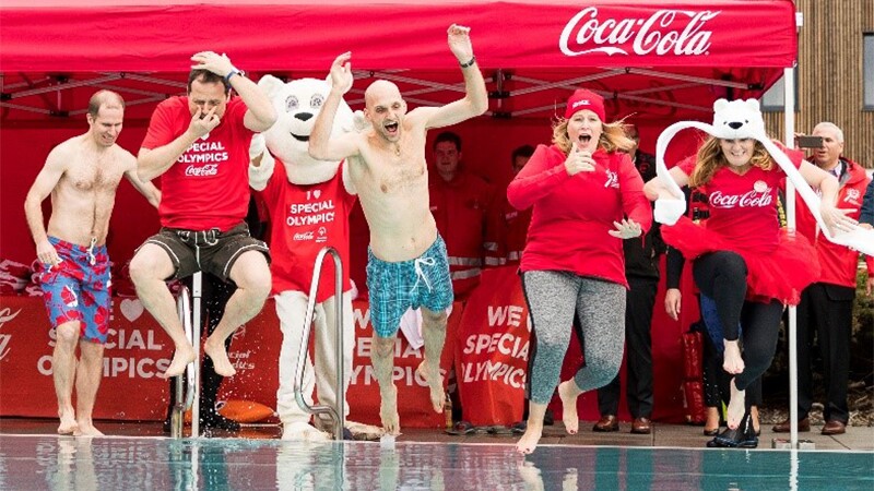 Four people jumping in the water at the Polar Plunge. The Coca Cola booth is in the background. 