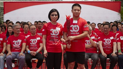 Man and woman holding a torch, an audience sits behind them and watches. 