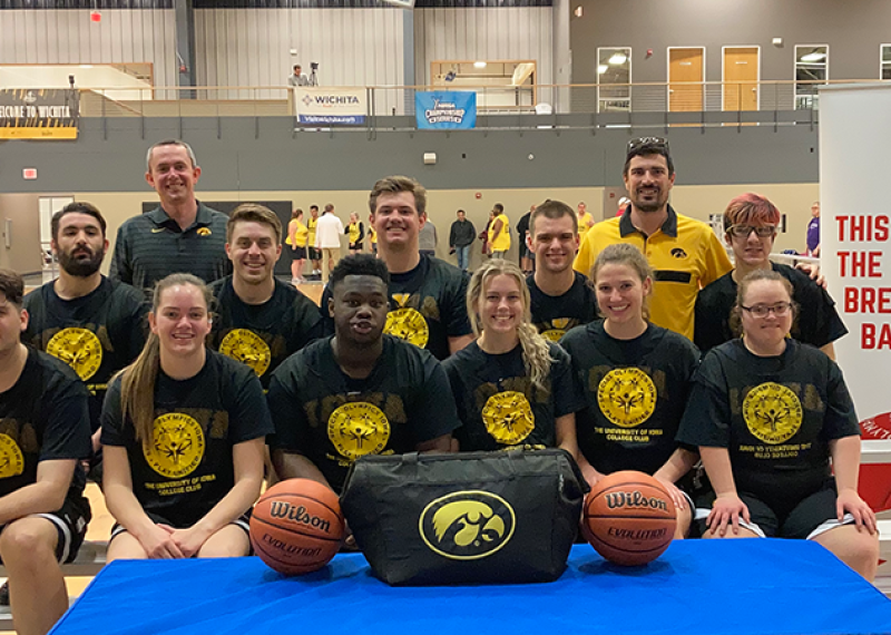 A group of men and women pose for a group photo in a basketball gym. They are wearing practice uniforms and smiling for the camera. 