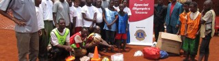 Group of people in 2 rows standing and one row crouching, outside next to Special Olympics Tanzania Lions Clubs signage.  