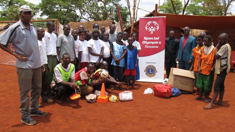 Group of people in 2 rows standing and one row crouching, outside next to Special Olympics Tanzania Lions Clubs signage.  