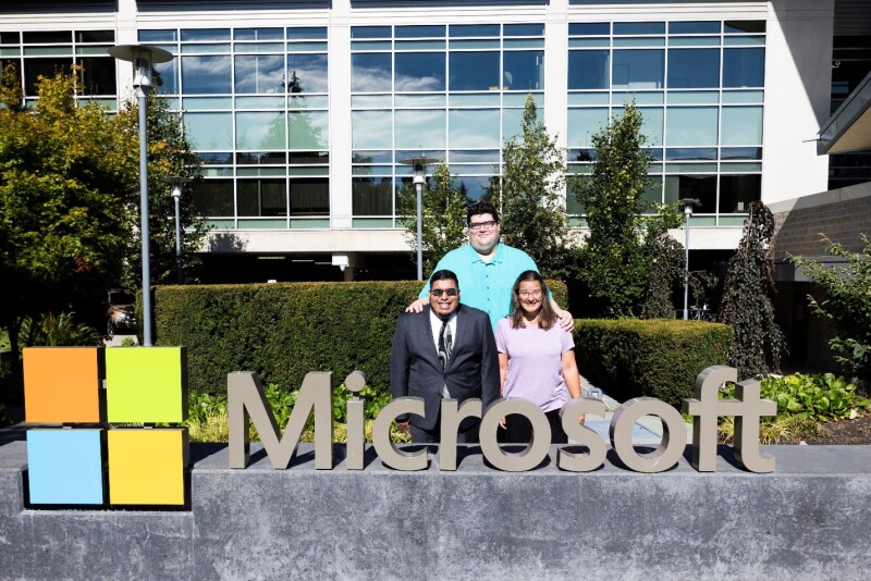 Three people standing behind a Microsoft sign at the Microsoft campus. 