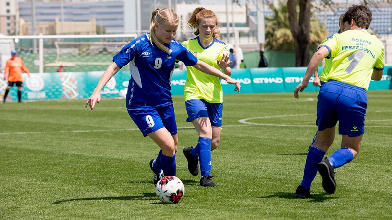 Three female footballers on the field. One girl is dribbling the ball as to opponents approach her. 