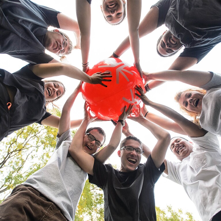 8 young adults standing in a circle holding a red ball looking down at the camera and smiling. 