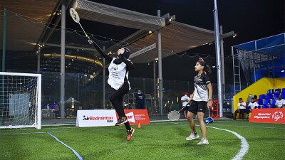 An athlete leaping in the air returning the Shuttlecock with her racket back over the net, with her partner also standing on the turf court next to her. 