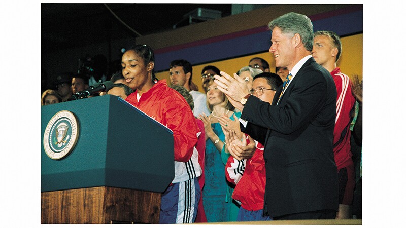 Loretta Clairborne speaking at a podium with the presidential seal next to President Bill Clinton and other Special Olympics supporters. 